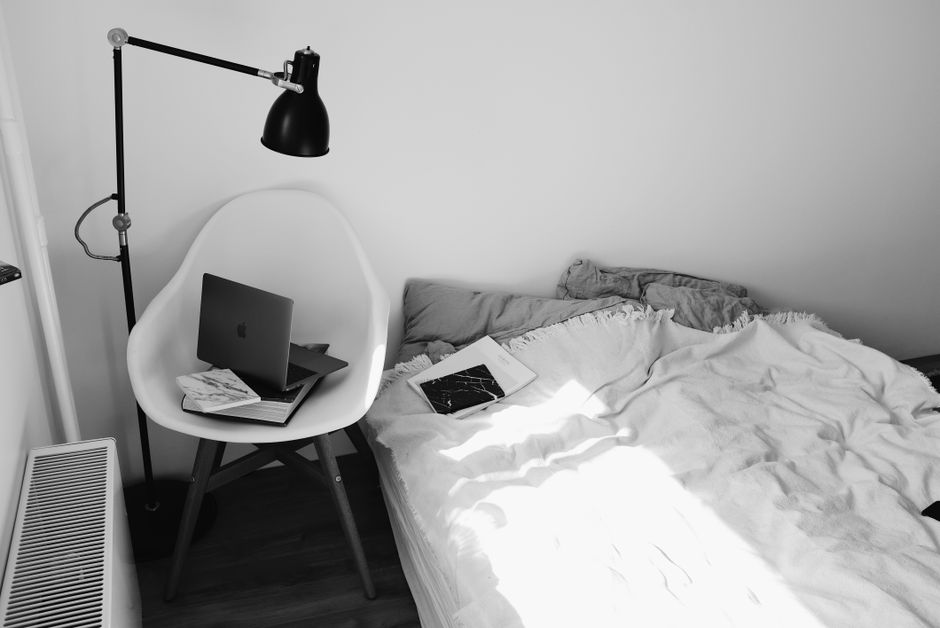 Imagem de um espaço de trabalho improvisado no quarto, com um notebook em uma cadeira ao lado da cama desfeita, ilustrando o perfil de alguém que trabalha ou estuda em ambientes informais.
