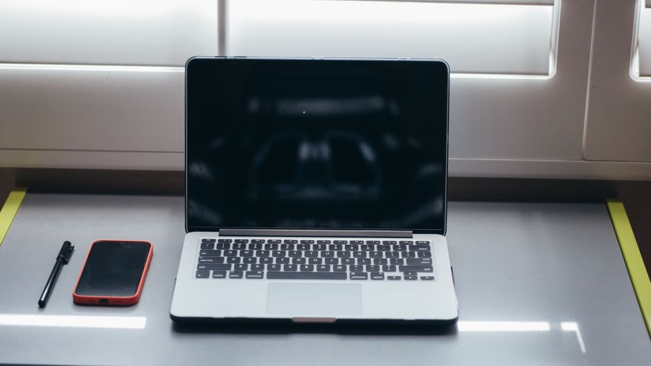 Imagem de espaço de trabalho com laptop, smartphone e caneta ao lado de uma janela, ideal para temas de produtividade e ambiente de trabalho remoto.
