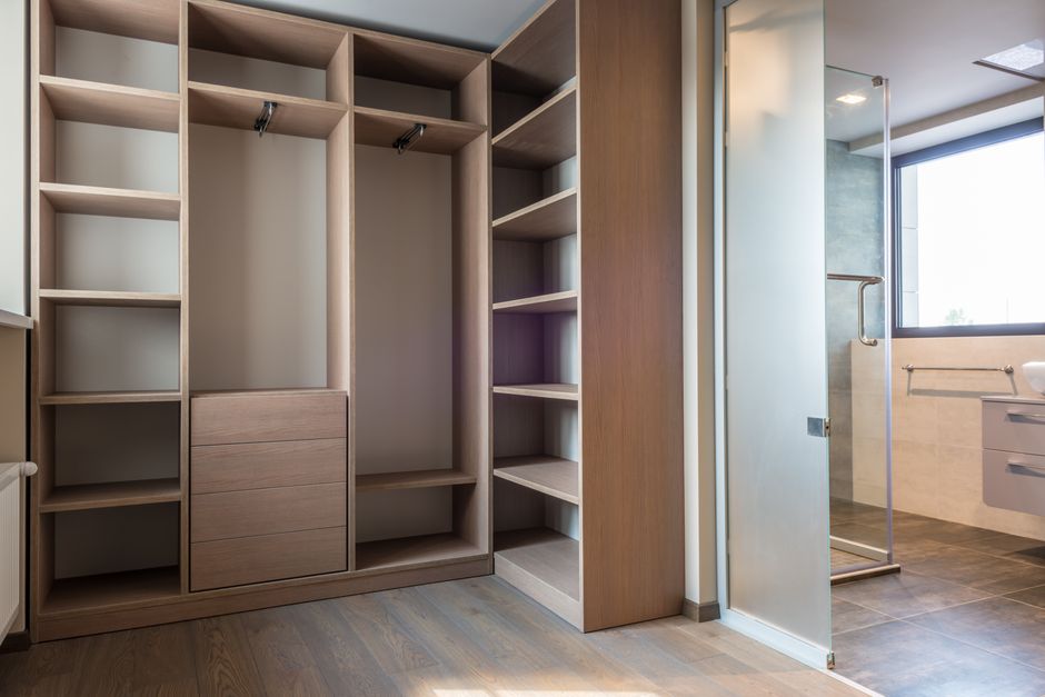 Imagem de um closet vazio com prateleiras de madeira ao lado de um banheiro com bancada e janela grande, em um ambiente moderno e bem iluminado.