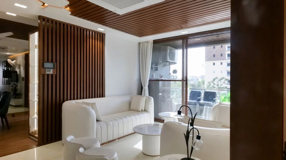 Sala de estar moderna com móveis brancos, detalhes em madeira e grande janela de vidro que proporciona iluminação natural e vista para um ambiente externo.