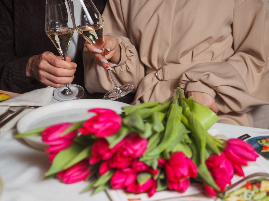 Casal brindando com taças de champagne, em um momento de celebração, à mesa com flores na decoração.