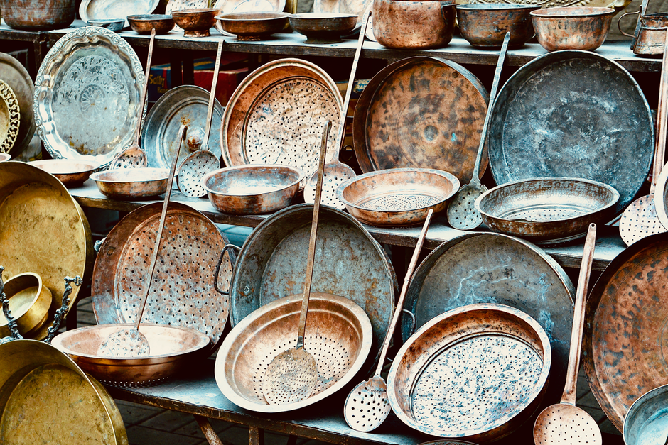 Imagem de utensílios de cobre e ferro usados em cozinhas tradicionais, formando uma coleção de panelas e espumadeiras expostas em uma loja ou feira de utensílios culinários.