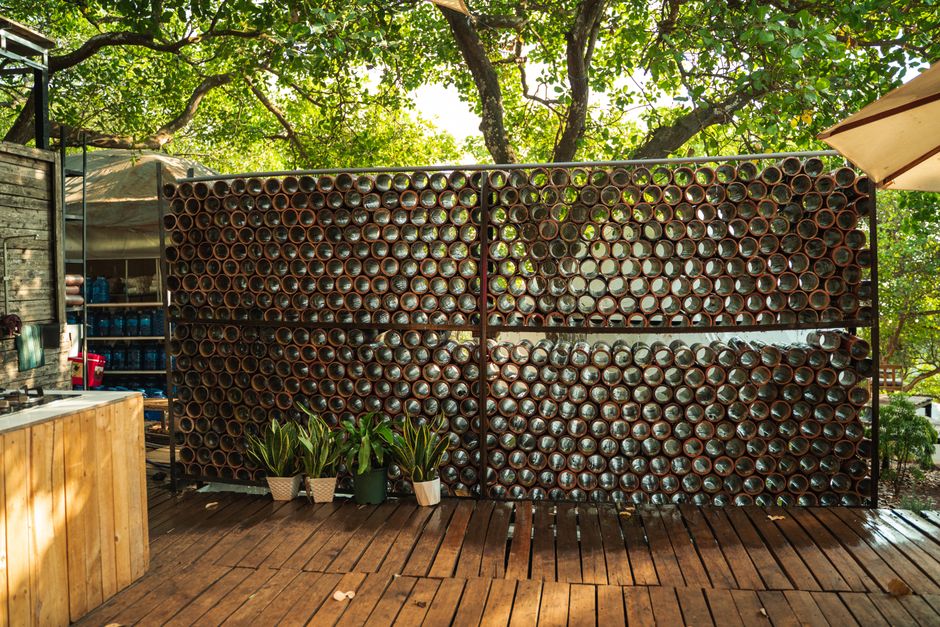 Imagem de uma parede de canos de metal empilhados em um ambiente ao ar livre, com plantas e sombra de árvores ao fundo, transmitindo uma sensação de sustentabilidade e design sustentável.