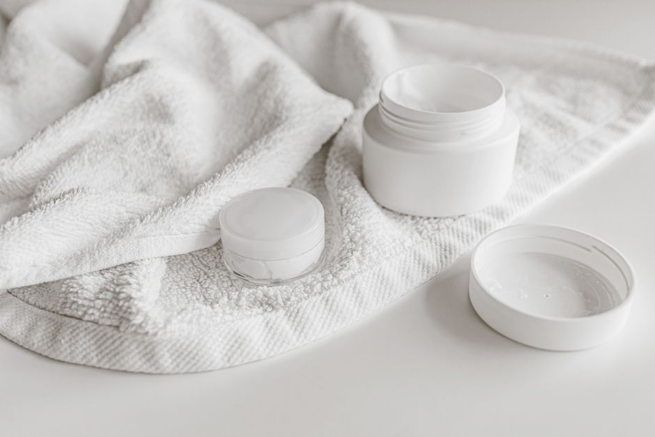 Imagem de produtos brancos de cuidados com a pele dispostos de forma minimalista sobre uma toalha branca, transmitindo sensação de limpeza e cuidados pessoais.