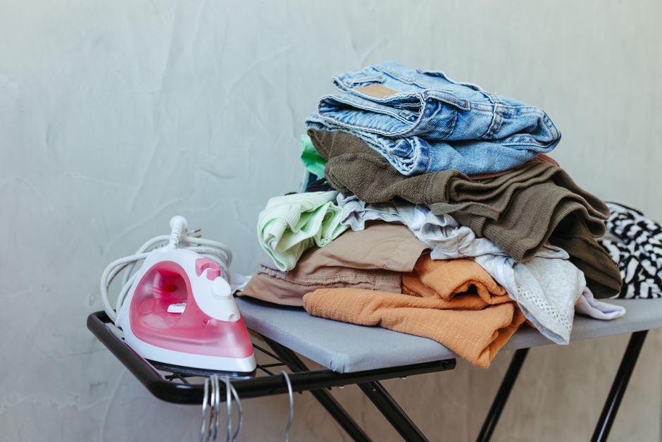 Imagem de uma pilha de roupas sujas sobre uma tábua de passar, ao lado de um ferro de passar roupas elétrico, sugerindo organização ou lavanderia doméstica.