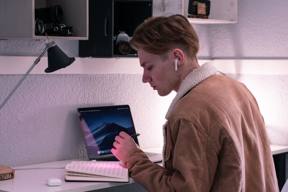 Jovem usando fones de ouvido, escrevendo em um caderno ao lado de um tablet, em um ambiente de estudo ou trabalho criativo.
