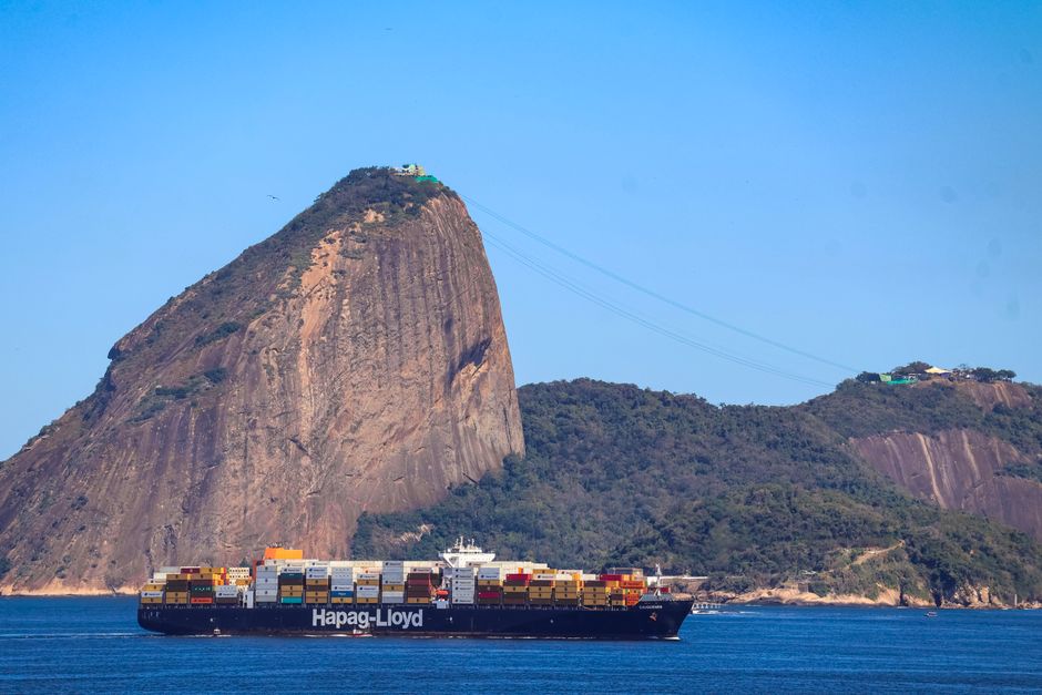 Vista de uma embarcação de transporte de cargas navegando próximo a uma paisagem de Montanha com teleférico e vegetação, na região do Rio de Janeiro.