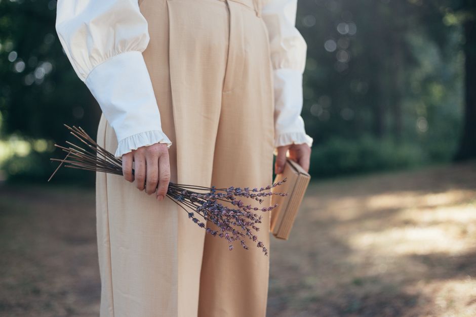 Pessoa vestida com roupa clássica, segurando um buquê de lavanda e um livro, em ambiente ao ar livre, simbolizando reflexão, nostalgia ou conexão com a natureza.