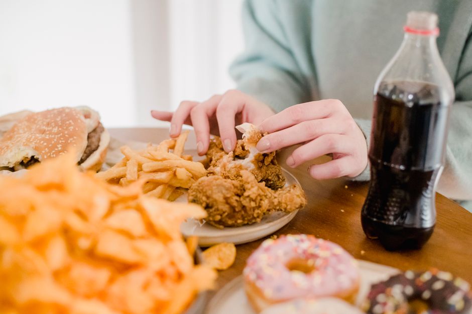 Pessoa comendo uma refeição rápida e comida de fast food, incluindo hambúrguer, batatas fritas e bolinho, ao lado de uma garrafa de refrigerante em um ambiente casual.