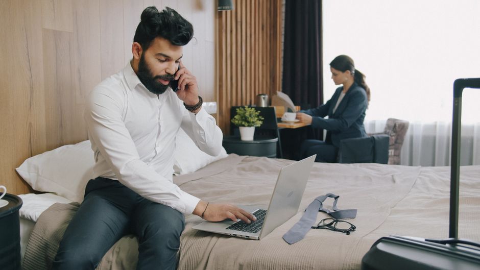 Homem trabalhando remotamente no hotel, realizando tarefas profissionais com laptop e telefone, em um ambiente de viagem de negócios.