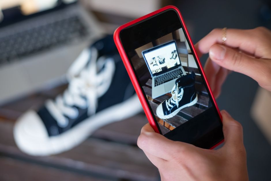 Pessoa tirando uma foto de um tênis e um laptop com uma câmera de smartphone, criando uma composição criativa para um blog de moda ou tecnologia.