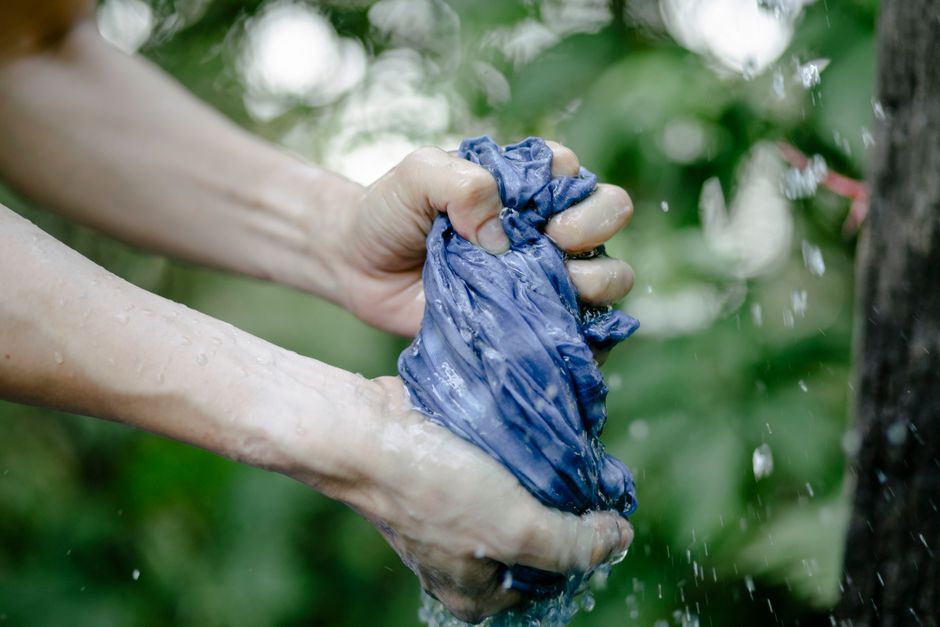 Pessoa lavando uma peça de roupa azul sob a água, destacando a atividade de lavagem manual ao ar livre.