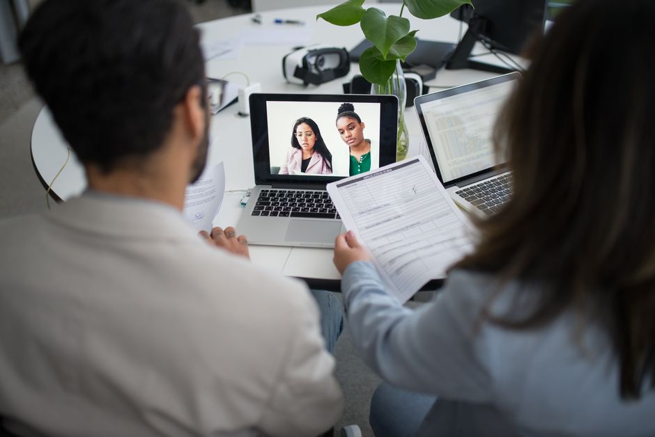 Reunião de equipe com duas pessoas assistindo a uma vídeo chamada, em ambiente de trabalho com laptops e documentos, focando na colaboração remota e comunicação empresarial.