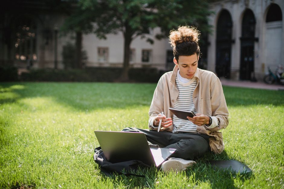 Jovem estudando e trabalhando ao ar livre em um parque, usando dispositivos eletrônicos e livros.