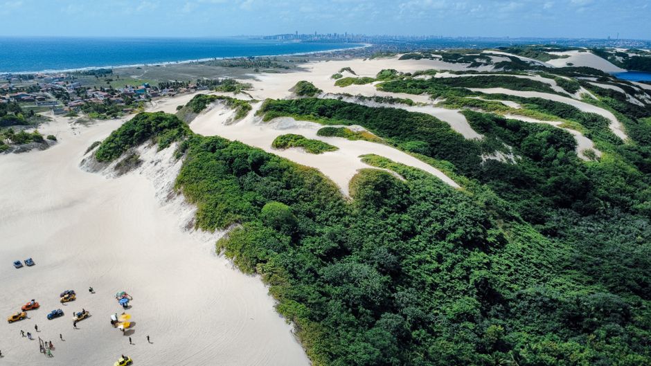 Imagem aérea de dunas de areia e vegetação costeira com o mar e cidade ao fundo, capturando a beleza natural e a preservação ambiental do litoral.