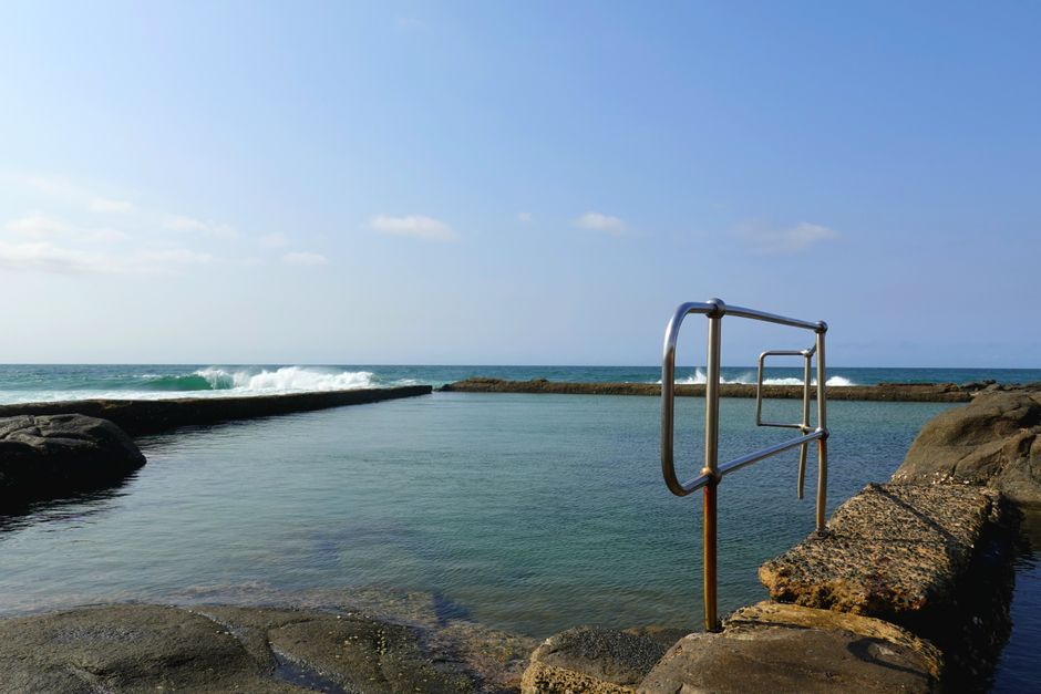 Uma piscina natural ao lado do mar com escada de metal para acesso ao banho de mar, em um cenário de praia ao ar livre.