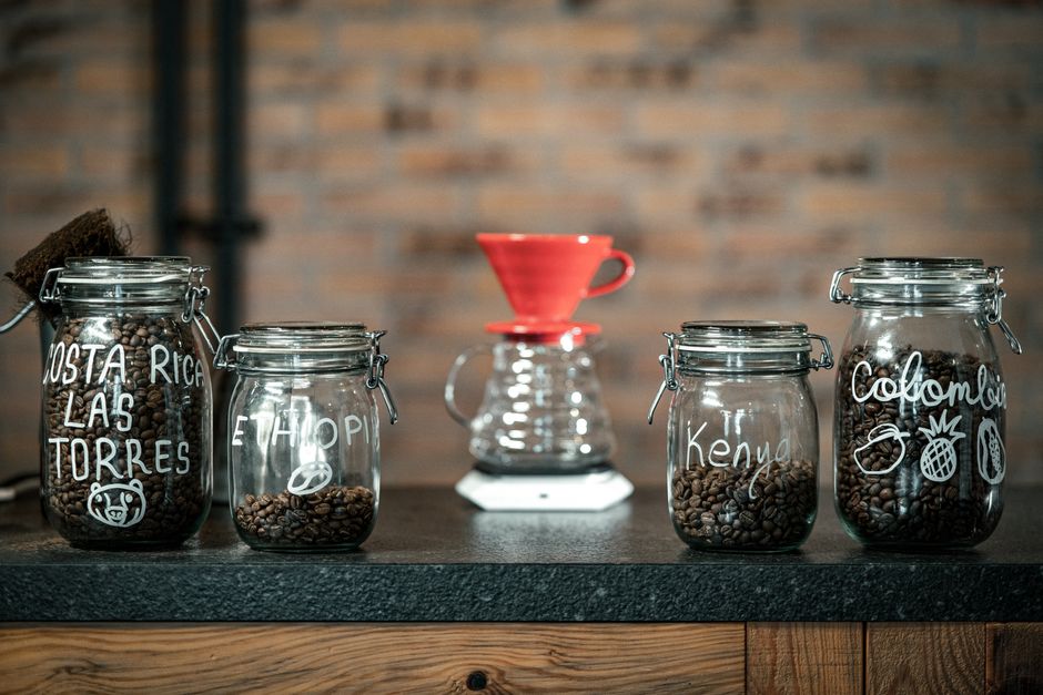 Imagem de diferentes tipos de grãos de café armazenados em potes de vidro, usados em uma cafeteria ou torrefadora artesanal.