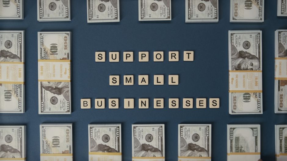 Imagem com destaque para a frase 'support small businesses' formada por blocos de letras, rodeada por notas de cem dólares, representando suporte a pequenos negócios e empreendedorismo.