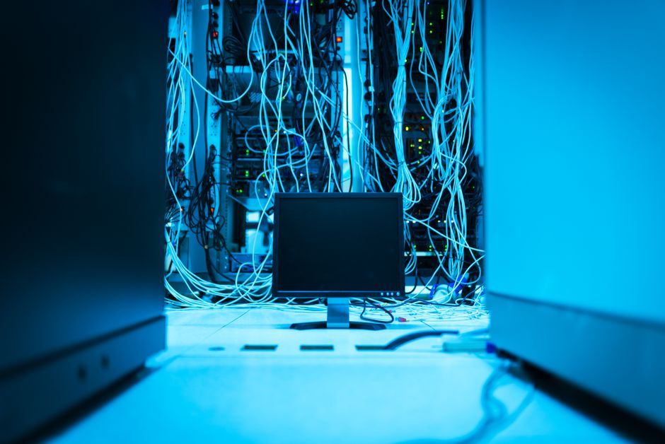 Imagem de um centro de dados com servidores conectados e uma tela de computador no ambiente tecnológico, simbolizando infraestrutura de TI e segurança de dados.