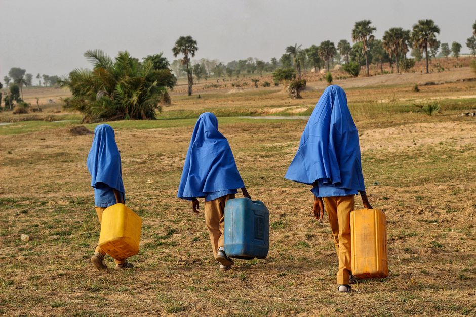 Três crianças com roupas azuis cobrindo suas cabeças carregando galões de água em uma paisagem rural seca, simbolizando desafios de acesso à água em regiões rurais.