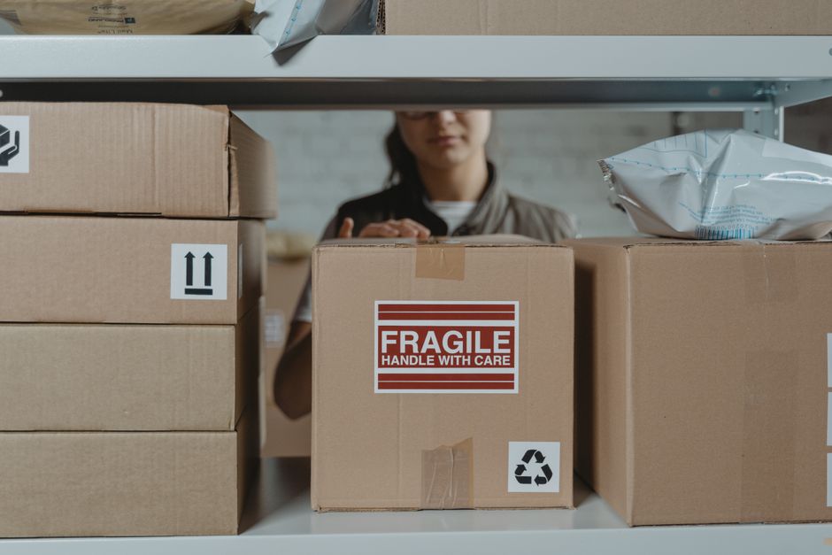 Pessoa organizando caixas de embalagem em um ambiente de armazenamento ou depósito, destacado pela etiqueta de fragilidade e símbolos de cuidado.