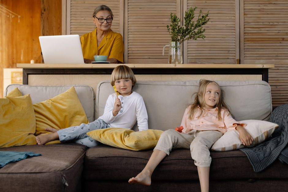 Uma mulher idosa trabalhando em home office com duas crianças calmamente descansando no sofá ao fundo.