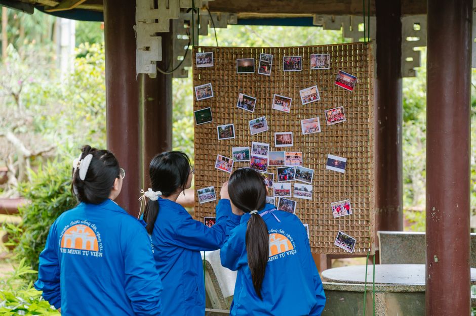 Jovens observando uma parede de fotos em ambiente ao ar livre, simbolizando memórias e conexão cultural.