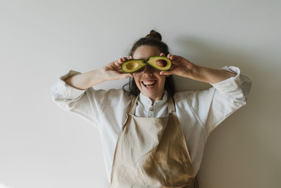 Mulher sorridente brincando com abacates na cabeça, transmitindo leveza e diversão na cozinha ou no cotidiano.