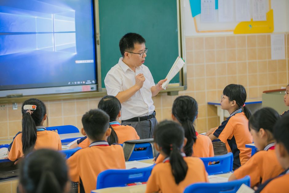 Professor lecionando para uma turma de estudantes em sala de aula, com quadro digital ao fundo.