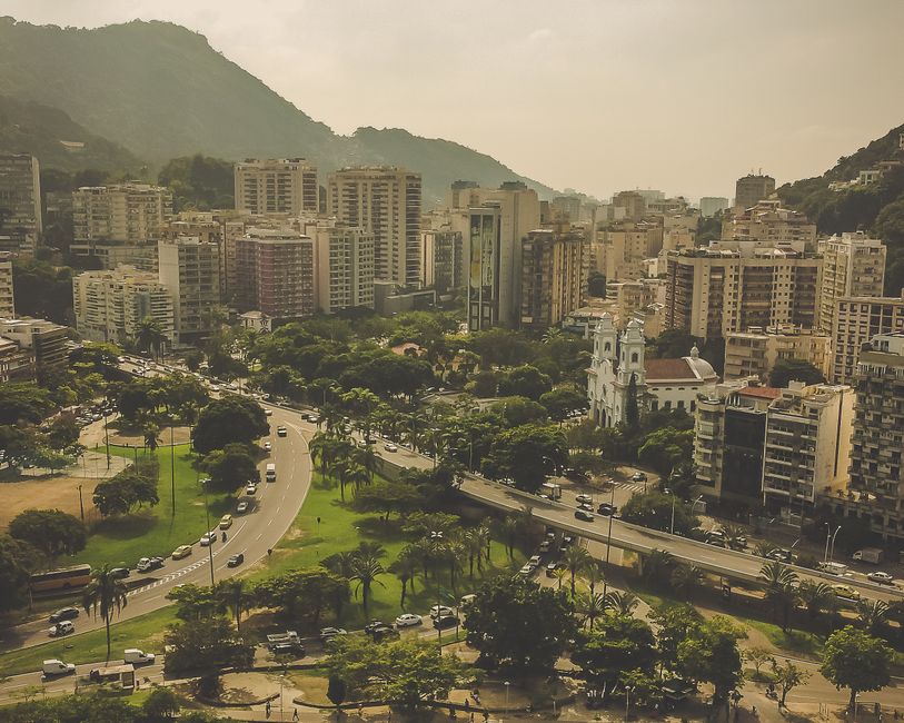 Uma vista panorâmica de uma cidade moderna com prédios altos, áreas verdes e uma igreja histórica no centro, com montanhas ao fundo.
