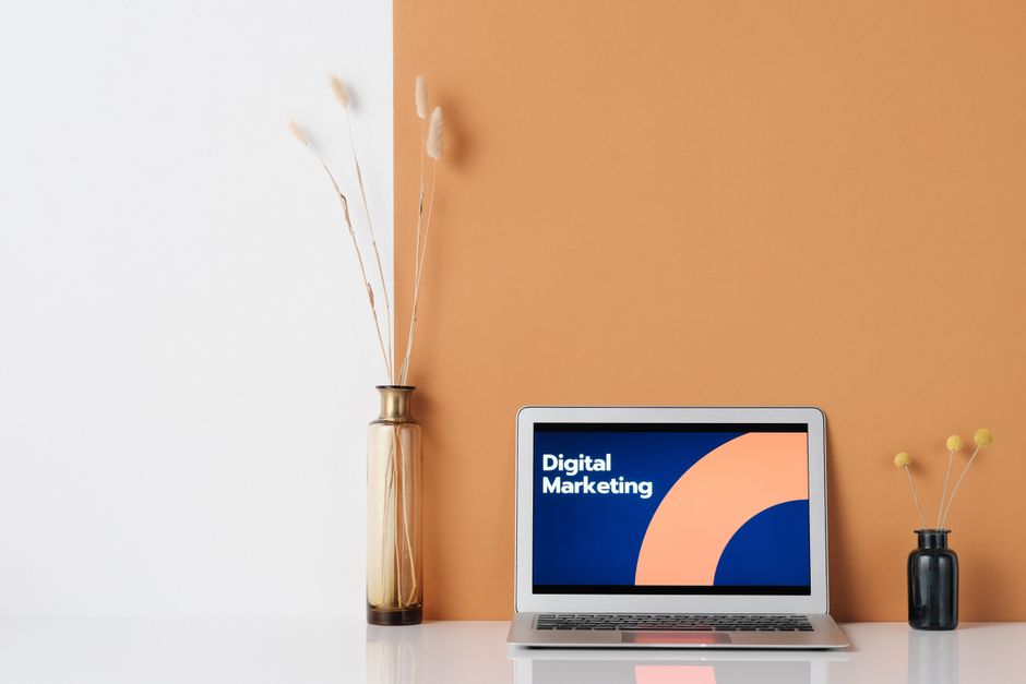 Imagem de um ambiente de trabalho com foco em marketing digital, apresentando um laptop com tela exibindo 'Digital Marketing' contra um fundo de parede dividida em cores neutras, decorado com vasos e plantas secas.