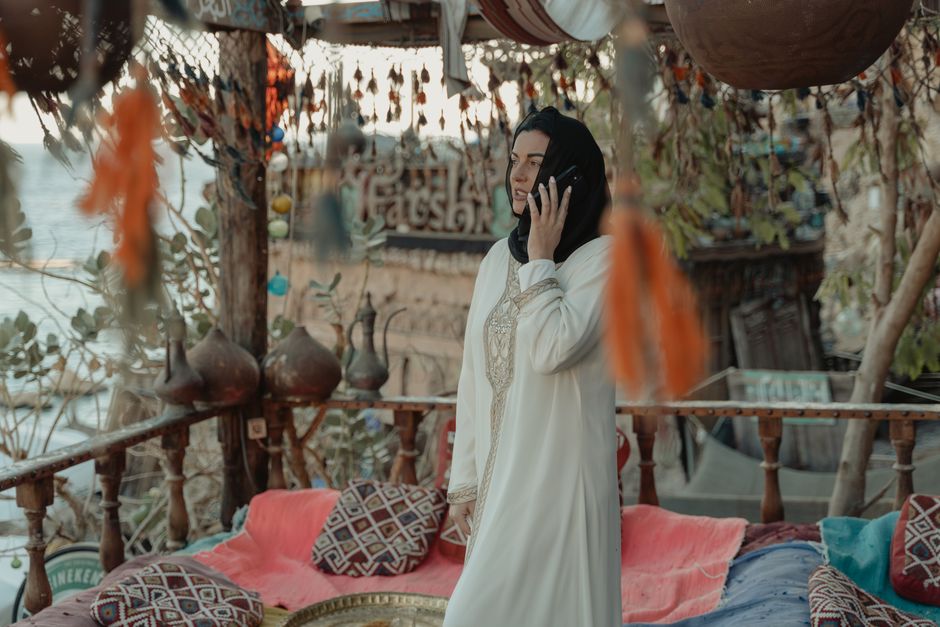Mulher de vestido branco e lenço preto falando ao telefone em ambiente ao ar livre decorado com objetos tradicionais e plantas, transmitindo tranquilidade e conexão cultural.