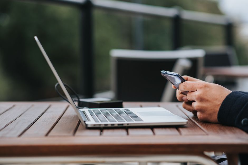 Pessoa usando smartphone ao lado de um laptop em ambiente externo, simbolizando trabalho remoto e conectividade profissional.