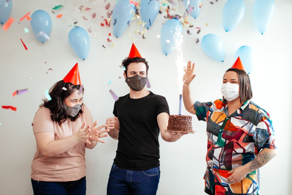 Celebrando aniversário de forma alegre, com balões, confetes e pessoas usando chapéus de festa e máscara de proteção.