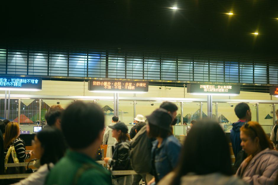 Imagem de pessoas esperando em fila em um terminal de transporte, com painéis de informações eletrônicos ao fundo, em ambiente urbano e movimentado.
