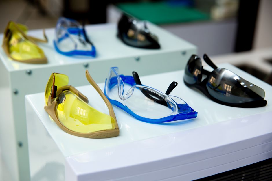 Vários modelos de óculos de proteção em cores distintas dispostos sobre displays, representando equipamentos de segurança para trabalhos industriais ou laboratoriais.
