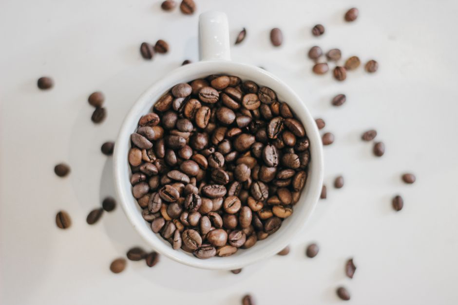 Café em grãos dentro de uma xícara branca, com alguns grãos espalhados ao redor, perfeito para ilustrar temas relacionados a cafés, produção de café ou cultura do café.