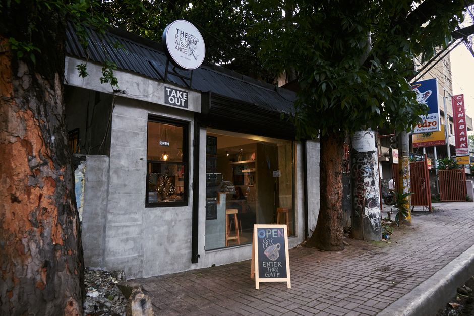 Pequeno estabelecimento de comida ou café visto na rua, com ambiente acolhedor e sinalização de aberto, rodeado por árvores e elementos urbanos.