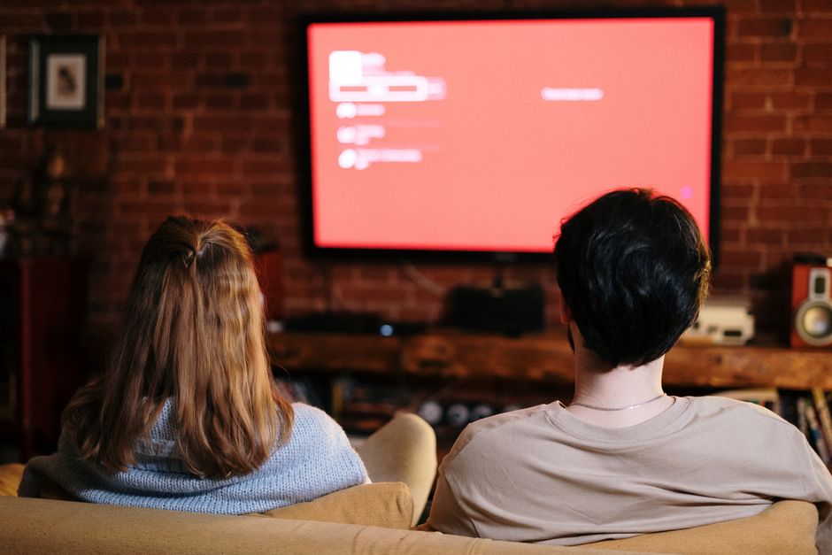 duas pessoas assistindo a uma apresentação ou filme em uma sala com parede de tijolos e televisão de tela grande