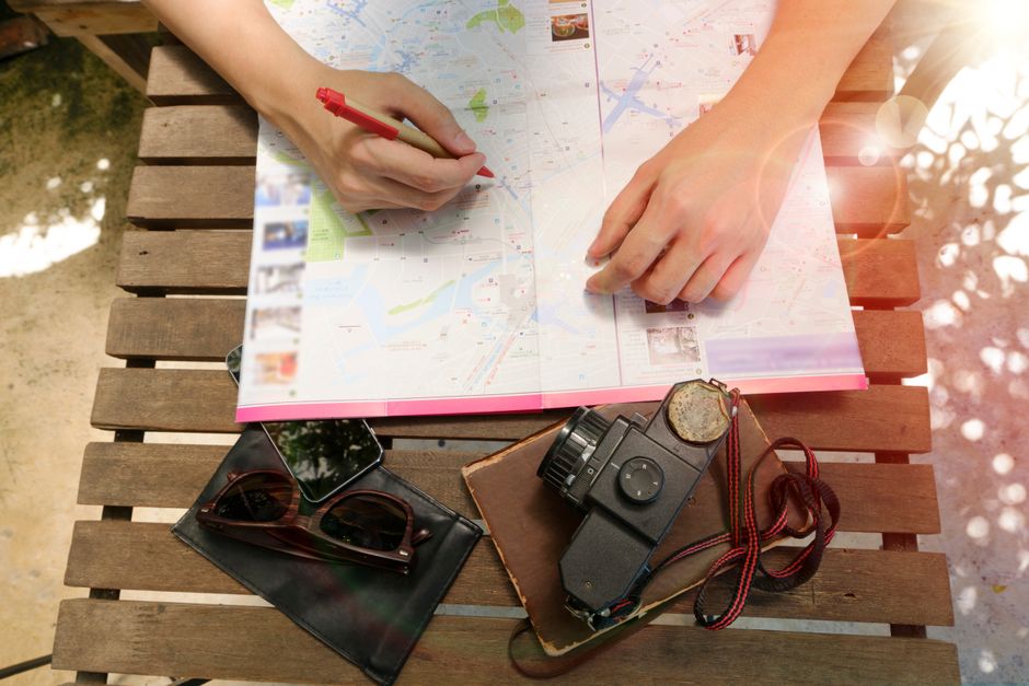 Duas pessoas planejando uma viagem ao ar livre, utilizando um mapa com uma câmera, óculos de sol e um celular sobre uma mesa de madeira.