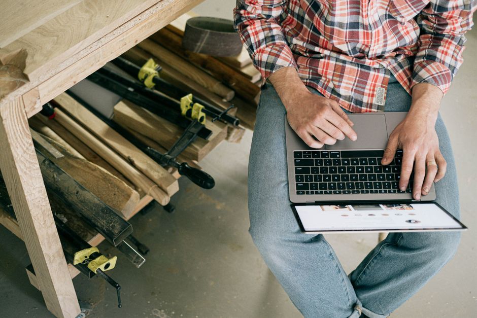 Homem utilizando um laptop em um ambiente de marcenaria ou madeira, sugerindo trabalho remoto ou pesquisa em um local de ofício.