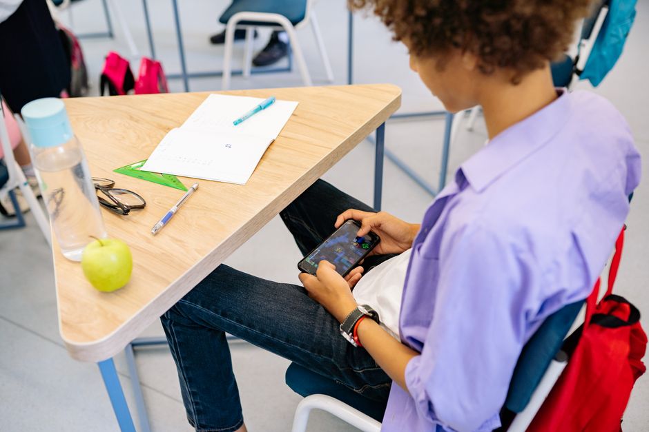 Pessoa estudando ou trabalhando em sala de aula, usando o celular com materiais de estudo na mesa ao seu redor.