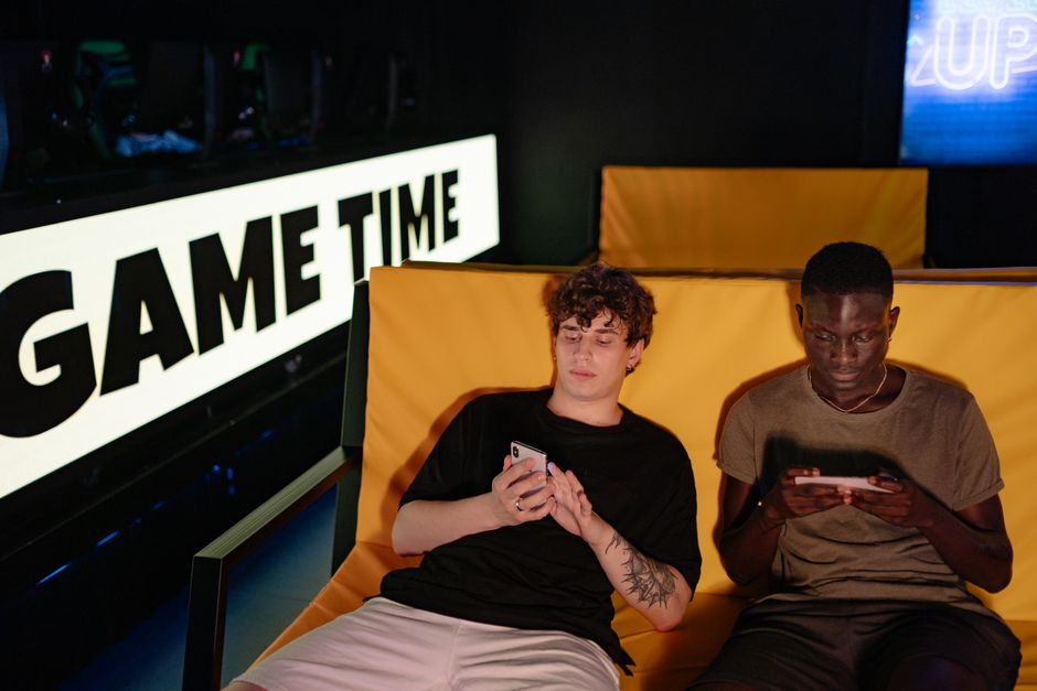 Dois jovens jogando em dispositivos móveis enquanto estão sentados em uma área de entretenimento com uma iluminação escura e um letreiro com a frase 'GAME TIME'.