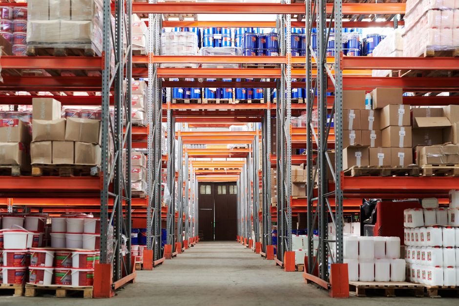 Imagem de um armazém com prateleiras cheias de caixas e, ao fundo, uma porta de saída, destacando a organização logística e o armazenamento de produtos.