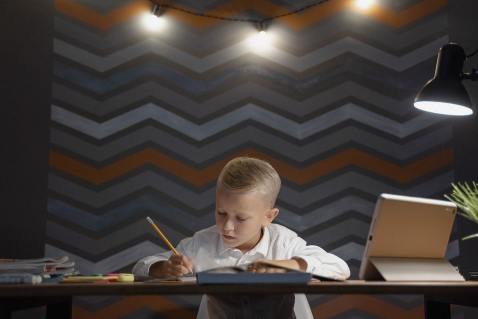 Criança estudando com foco em um ambiente de trabalho ou estudo noturno, com iluminação direcionada e elementos de organização ao redor.