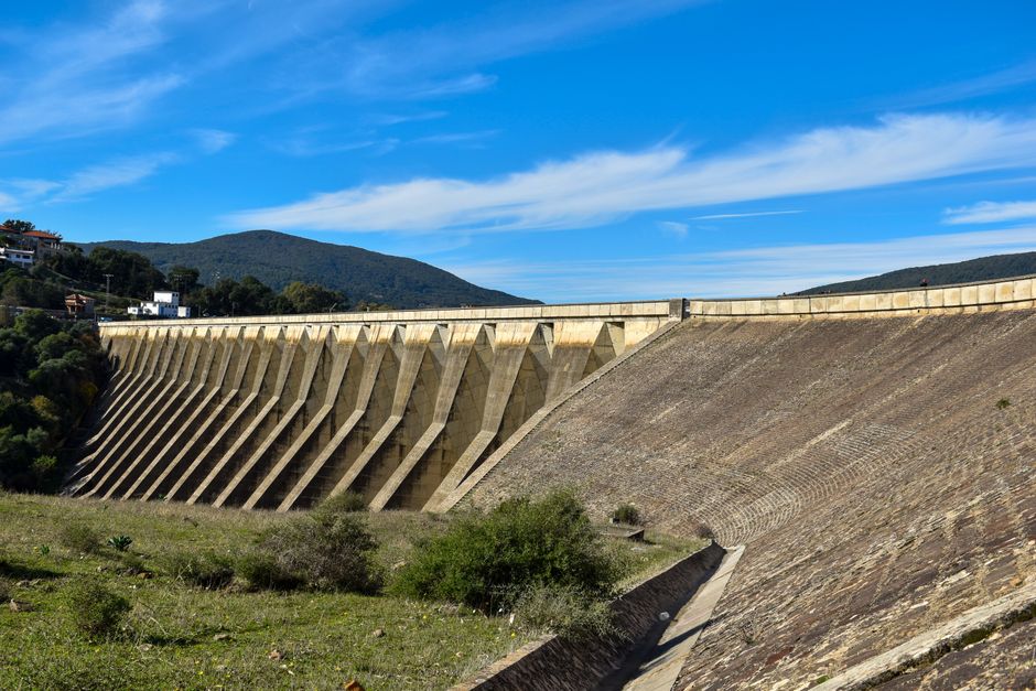 Imagem de uma barragem de contenção de água em um ambiente natural, com montanhas ao fundo e céu azul claro.