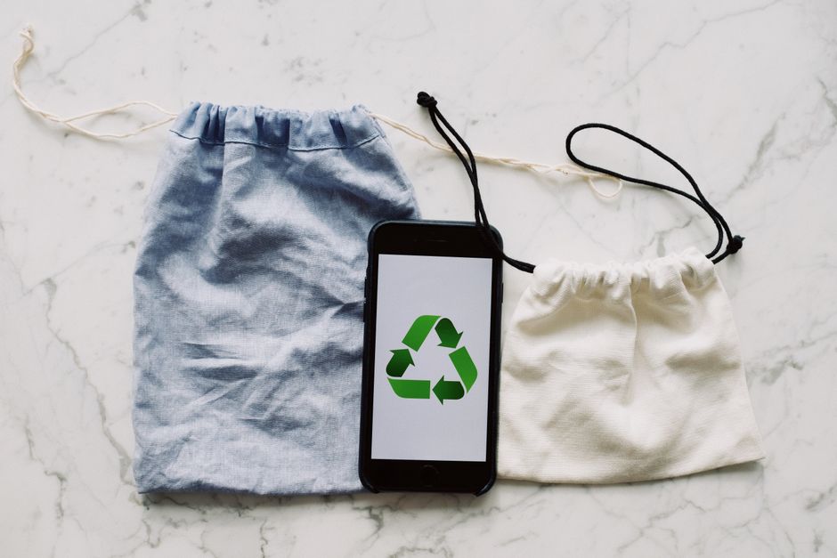 Imagem de um celular com símbolo de reciclagem na tela, acompanhado de dois saquinhos de tecido, sugerindo sustentabilidade e consumo consciente.