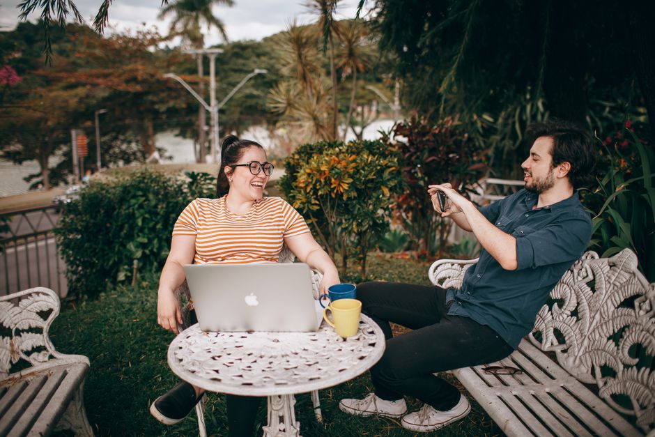 Duas pessoas sorrindo e conversando ao ar livre, em um ambiente tranquilo com vegetação e uma mesa de jardim.