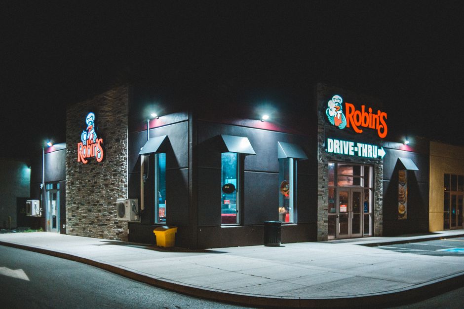 Ilustração de uma loja de fast food com destaque para o restaurante Robin's em um ambiente noturno, com iluminação externa e sinalização visível.