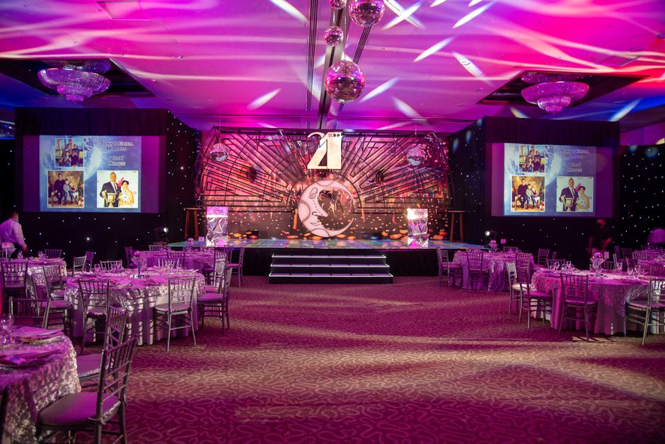 Sala de evento decorada com iluminação rosa e palco preparado para uma cerimônia ou celebração, com destaque para elementos glamourosos e sofisticados.